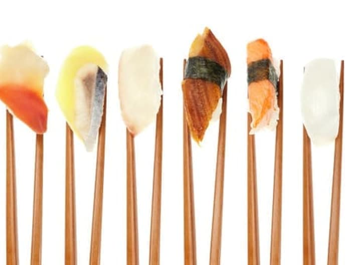 A pair of chopsticks grips but doesn't crush food - 17 different types of sushi being held up in a row with wooden chopsticks isolated on white.